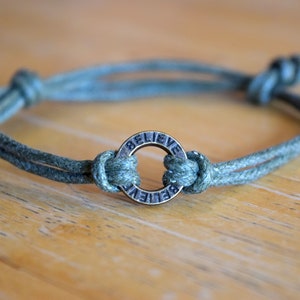 May include: A green cord bracelet with a metal charm that says "Believe" in a circle.