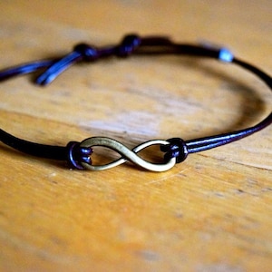 Könnte beinhalten: Ein braunes Lederarmband mit einem Unendlichkeits-Anhänger. Der Anhänger ist aus Messing und hat eine goldene Oberfläche.