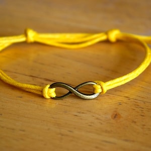 Puede incluir: Una pulsera de cuerda amarilla con un dije de símbolo de infinito. El dije está hecho de metal y tiene un acabado dorado.