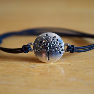 May include: A black string bracelet with a silver charm featuring a tree of life design.