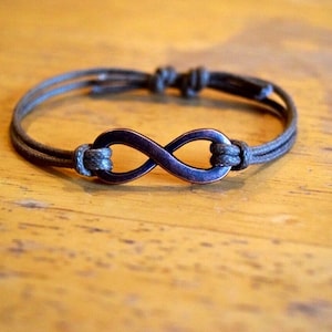 May include: A brown cord bracelet with an infinity symbol charm. The charm is made of metal and has a dark, almost black, finish.