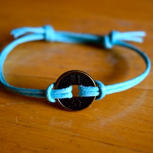 Könnte beinhalten: Ein türkisfarbenes Schnurarmband mit einem runden, goldfarbenen Anhänger mit chinesischen Schriftzeichen. Der Anhänger ist mit einem Knoten an der Schnur befestigt.