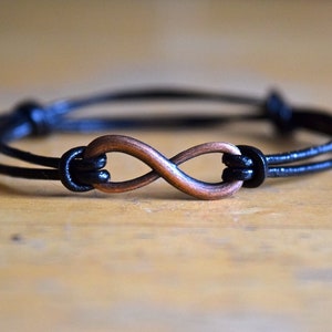 Op de afbeelding: Een zwart leren armband met een koperen oneindigheidssymbool bedel.