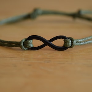 May include: An olive green cord bracelet with a black infinity symbol charm. The bracelet has an adjustable design. The infinity symbol is a popular symbol of eternity and love. The cord is approximately 1 mm thick.