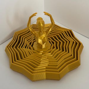 May include: A gold-colored 3D printed sculpture featuring a central figure of a dancer with arms raised above the head, surrounded by a series of concentric, tiered, octagonal-shaped rings that create a layered, expanding base structure.