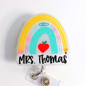 May include: A white glitter badge holder with a rainbow design featuring a red apple, paper clips, and a pencil. The text "MRS. THOMAS" is printed in black on the badge holder.