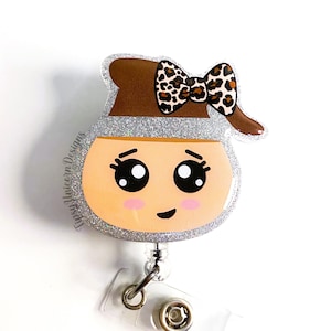 May include: A silver glitter badge holder with a brown and orange cartoon face with a black and brown leopard print bow. The badge holder has a clear plastic clip.