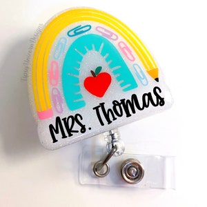 May include: A white glitter rainbow with yellow, pink, and blue accents. The rainbow has paper clips and a pencil on it. There is a red apple in the center of the rainbow. The text "Mrs. Thomas" is written in black on the rainbow. The rainbow is attached to a clear plastic badge holder with a silver retractable reel.