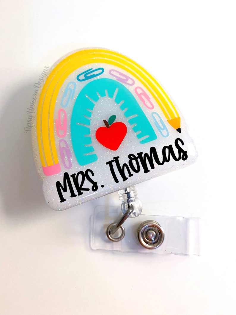 May include: A white glitter rainbow with yellow, pink, and blue accents. The rainbow has paper clips and a pencil on it. There is a red apple in the center of the rainbow. The text "Mrs. Thomas" is written in black on the rainbow. The rainbow is attached to a clear plastic badge holder with a silver retractable reel.
