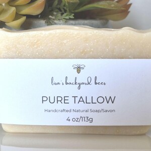 May include: A rectangular bar of off-white soap wrapped in a white paper label. The label features a bee icon, the text 'lisa's backyard bees', 'PURE TALLOW', 'Handcrafted Natural Soap/Savon', and the weight '4 oz/113g' printed in black ink.