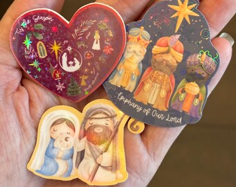 May include: A collection of Christmas-themed stickers. One is heart-shaped with text that reads "Tis the Season" and features a nativity scene. Another depicts the three wise men, and the third shows Mary, Joseph, and baby Jesus.
