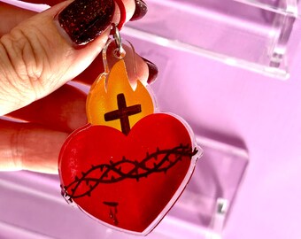 May include: A red heart-shaped keychain with a black crown of thorns and a cross. The keychain has a red clasp and a flame design above the heart. The background is a light purple color.