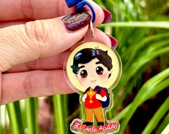 May include: A clear acrylic keychain featuring a cartoon illustration of a young boy with black hair, wearing a red shirt, backpack, and holding a cross. The keychain has a blue clasp and the text "St. Carlo Acutis".