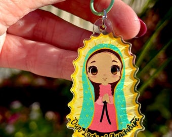 May include: A clear acrylic keychain featuring a cartoon illustration of Our Lady of Guadalupe. The image has a yellow halo, a teal veil, and a pink dress. The keychain has a green clasp.