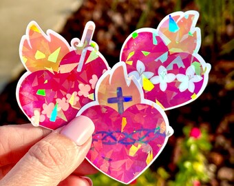 May include: Three iridescent holographic stickers of pink hearts with white flowers and a cross. The hearts are outlined in white and have a glittery finish.