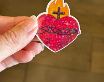 May include: A red heart with a crown of thorns and a cross on top, surrounded by a halo of flames. The heart is covered in glitter and has a holographic effect.