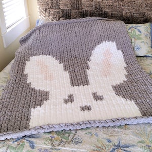 May include: A gray and white knitted throw blanket with a white bunny rabbit design. The blanket is on a bed with a floral patterned bedspread.