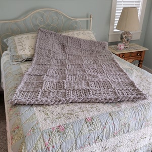 Op de afbeelding: Een grijze gebreide deken met een textuurpatroon is gedrapeerd over een bed met een quilt met bloemenpatroon. De deken is gemaakt van dik garen en voelt zacht en comfortabel aan.