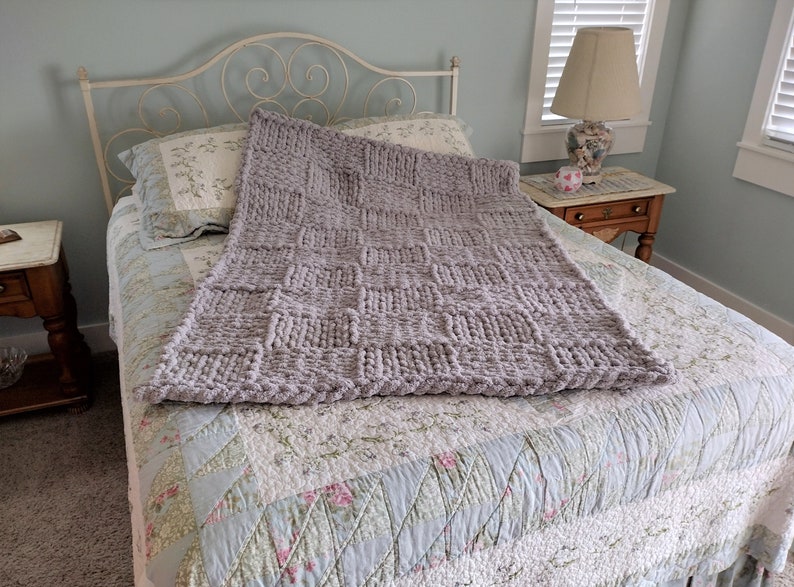 Op de afbeelding: Een grijze gebreide deken met een textuurpatroon is gedrapeerd over een bed met een quilt met bloemenpatroon. De deken is gemaakt van dik garen en voelt zacht en comfortabel aan.