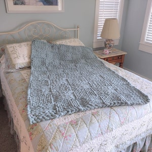 Op de afbeelding: Een lichtblauwe gebreide deken met een textuurpatroon, gedrapeerd over een bed met een bedsprei met bloemenpatroon.