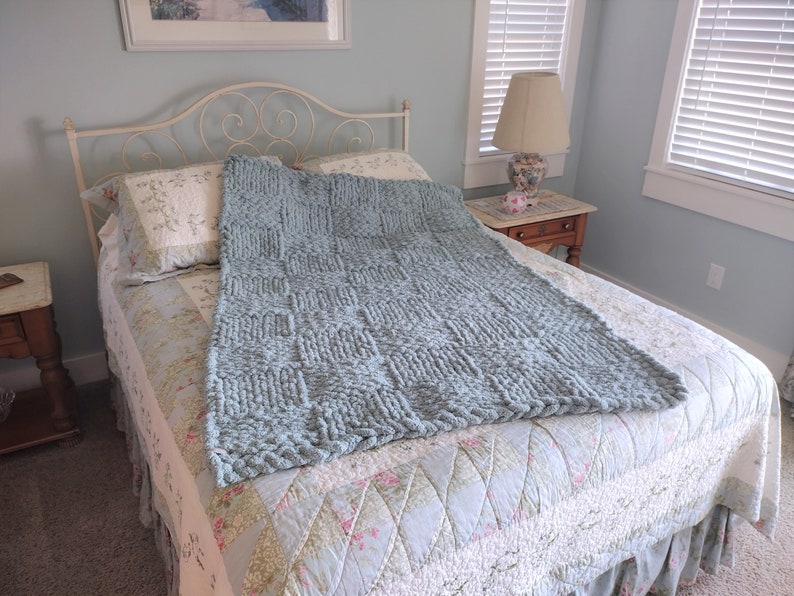 Op de afbeelding: Een lichtblauwe gebreide deken met een textuurpatroon, gedrapeerd over een bed met een bedsprei met bloemenpatroon.