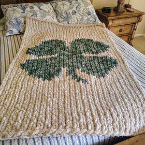 May include: A large, cream-colored knitted blanket with a green four-leaf clover design. The blanket is on a bed with blue and white striped bedding.
