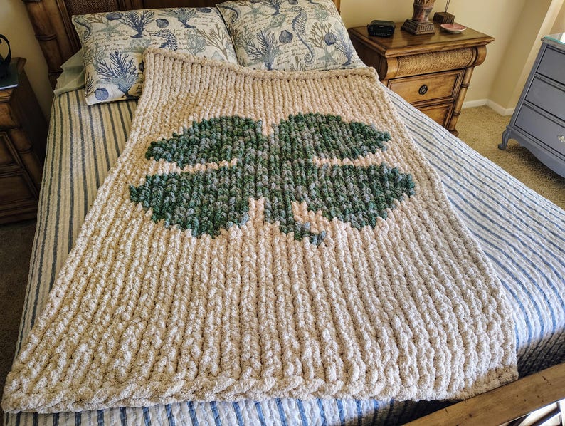 May include: A large, cream-colored knitted blanket with a green four-leaf clover design. The blanket is on a bed with blue and white striped bedding.