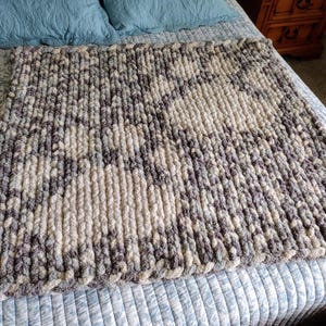 Puede incluir: Una manta de punto grueso en un esquema de colores gris y blanco. La manta está hecha con hilo grueso y tiene un aspecto texturizado y tejido.