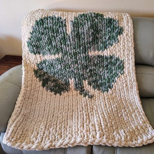 May include: A cream-colored knitted throw blanket with a large green four-leaf clover design.