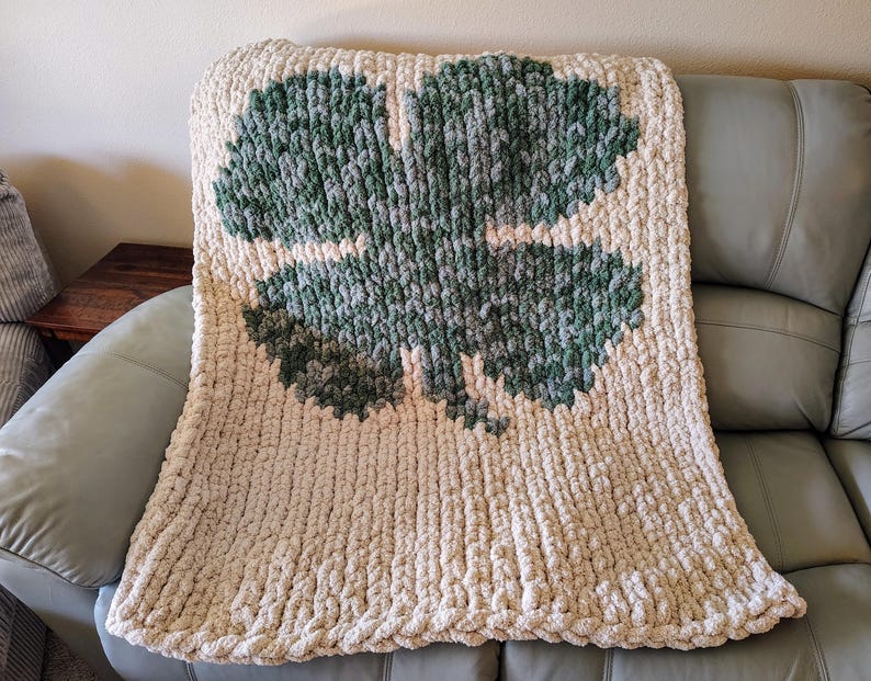 May include: A cream-colored knitted throw blanket with a large green four-leaf clover design.