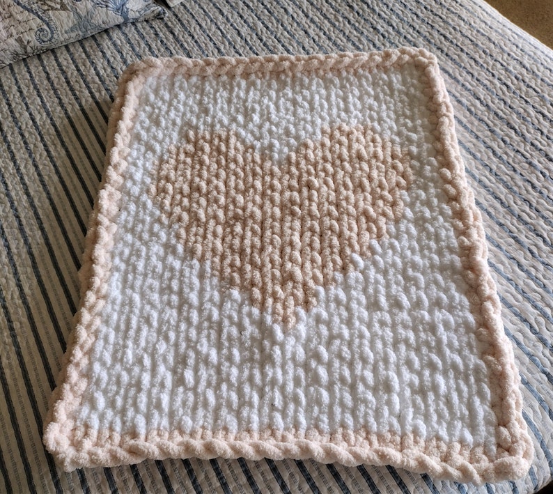 May include: A white and blush pink crocheted baby blanket with a heart design.