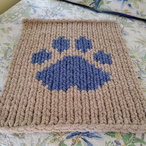 May include: A tan knitted throw blanket with a blue paw print design. The blanket is made of chunky yarn and has a soft, fluffy texture.