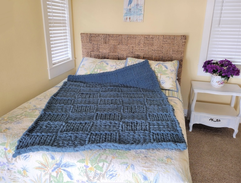 Op de afbeelding: Een blauwe gebreide deken met een geweven patroon op een bed met een rieten hoofdeinde. Het bed heeft een dekbedovertrek met een bloemenpatroon en twee kussens. Een klein wit nachtkastje met een vaas met paarse bloemen staat op de achtergrond.