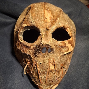 May include: A weathered, handmade mask with a textured, brown surface. The mask features large, dark eye holes, a small nose opening, and stitched details. The overall design suggests a rustic or antique aesthetic, suitable for costume or display.