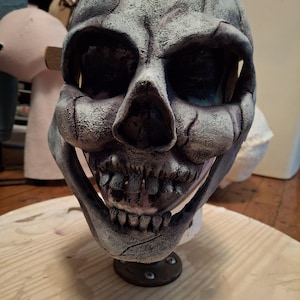 Puede incluir: Una máscara de calavera detallada, pintada a mano, con un acabado texturizado gris y blanco. La máscara tiene una boca ancha abierta con dientes visibles.
