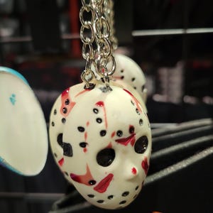 May include: A white plastic keychain featuring a red and black hockey mask design. The mask has a black strap and a white face with red blood splatters.