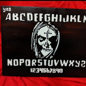 May include: A black wooden Ouija board with white lettering and numbers. The board features a white image of a skull with a cross on its forehead. The words "yes" and "no" are written in the top corners of the board.