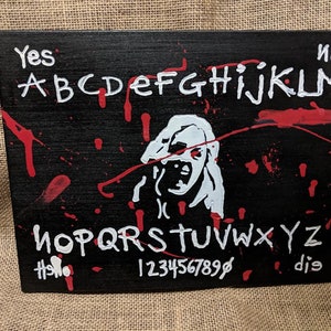 May include: A black wooden Ouija board with red paint splatters. The board features a white silhouette of a screaming face, the letters of the alphabet, numbers 1-9 and 0, and the words "yes", "no", "hello", and "goodbye".