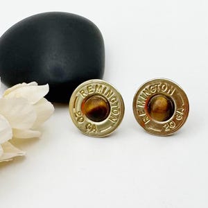 May include: A pair of circular stud earrings crafted from gold-toned metal Remington 20 GA shotgun shell casings. Each earring features a polished, round, brown and gold tiger's eye gemstone set in the center of the metallic base.