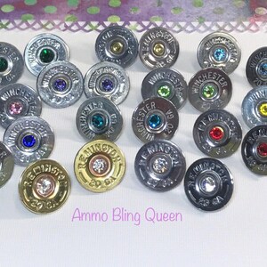 May include: A collection of 20 gauge shotgun shell earrings with various colored rhinestones. The earrings are silver and gold in color. The text "Ammo Bling Queen" is visible in the image.