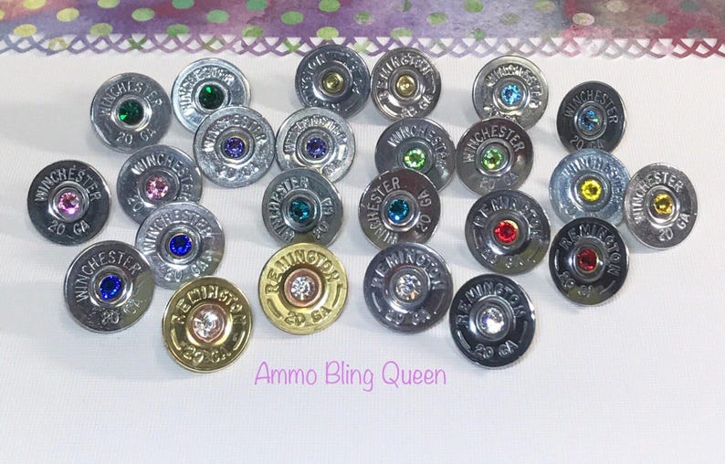 May include: A collection of 20 gauge shotgun shell earrings with various colored rhinestones. The earrings are silver and gold in color. The text "Ammo Bling Queen" is visible in the image.