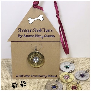 May include: A gold shotgun shell charm with a clear rhinestone, hanging from a pink ribbon. The charm is labeled "Winchester 12 GA" and "AA". The charm is displayed on a brown paper house-shaped tag with the text "Shotgun Shell Charm By Ammo Bling Queen" and "A Gift For Your Furry Friend".