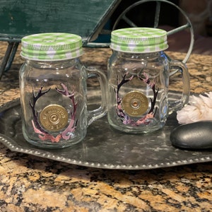 May include: Two glass salt and pepper shakers with green and white checkered lids. The shakers are decorated with a hunting theme, featuring a shotgun shell with antlers and camouflage print.