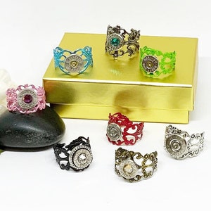 May include: A set of seven rings made from bullet casings and decorative metal filigree. The rings are in various colors, including pink, green, red, blue, and black. Each ring features a different colored gemstone.