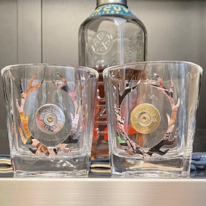 May include: Two clear glass tumblers with a camouflage pattern and a shotgun shell design. The tumblers are on a shelf with a bottle of whiskey in the background.