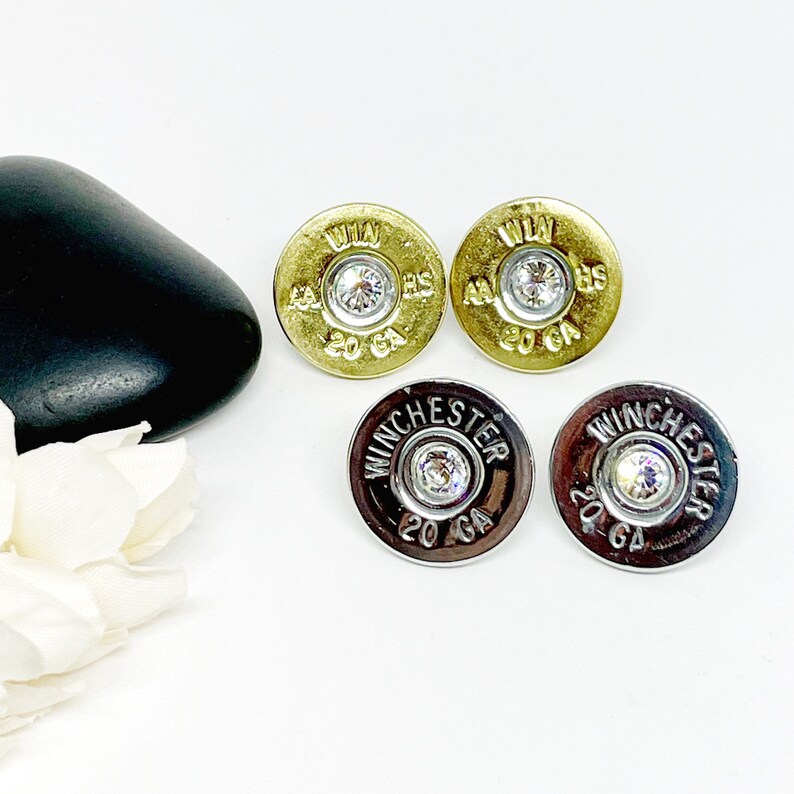 May include: A pair of earrings made from 20 gauge shotgun shells with a clear rhinestone in the center. The shells are gold and silver.
