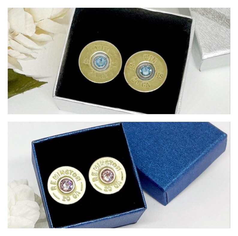 May include: Two pairs of gold-colored shotgun shell earrings with blue and pink gemstones. The earrings are in their boxes, one with a blue velvet interior and the other with a black velvet interior. The boxes are both closed.