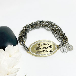 May include: Gold charm bracelet with a gold oval charm that says "She leaves a little sparkle wherever she goes" with rhinestones around the edge. The bracelet has multiple silver and gold chains and a silver charm with the letter "D".
