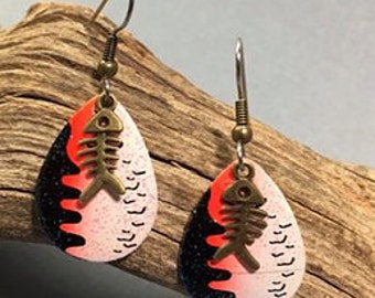 May include: A pair of dangle earrings with a teardrop-shaped design. The earrings are black and white with a red drip effect and a gold fishbone charm.
