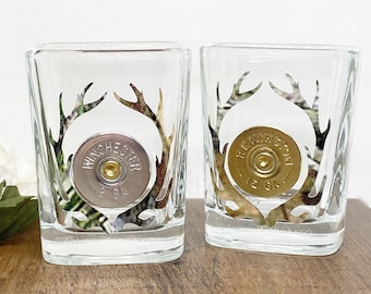 May include: Two clear shot glasses with a deer antler design and a bullet casing in the center. One glass has "Winchester 12 GA" and the other has "Remington 12 GA" printed on the casing.